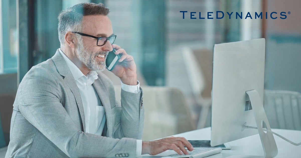 Businessman looking at a computer monitor while speaking into a smartphone - TeleDynamics blog
